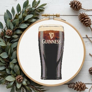 Pu&ograve; includere: Un ricamo a punto croce di una pinta di Guinness, con l'iconico logo e una schiuma cremosa. L'opera &egrave; esposta all'interno di un telaio da ricamo in legno, circondata da verde e pigne, creando un'estetica rustica.