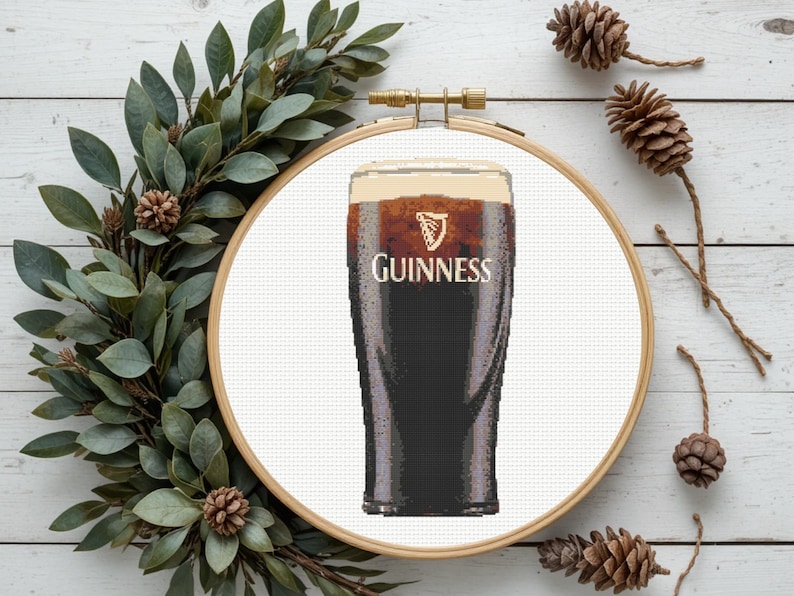 Pu&ograve; includere: Un ricamo a punto croce di una pinta di Guinness, con l'iconico logo e una schiuma cremosa. L'opera &egrave; esposta all'interno di un telaio da ricamo in legno, circondata da verde e pigne, creando un'estetica rustica.