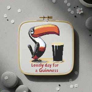 May include: Cross-stitch embroidery in a square wooden hoop. The design features a cartoon toucan with a large orange beak and a pint of Guinness. Text reads "Lovely day for a Guinness."