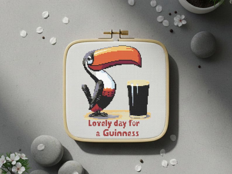May include: Cross-stitch embroidery in a square wooden hoop. The design features a cartoon toucan with a large orange beak and a pint of Guinness. Text reads "Lovely day for a Guinness."