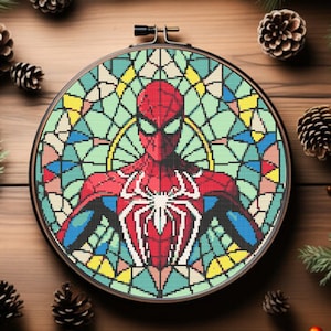 May include: A round embroidery hoop with a colorful stained glass design featuring a red and blue superhero with a spider web on their chest. The design is surrounded by a green, blue, and yellow stained glass pattern.