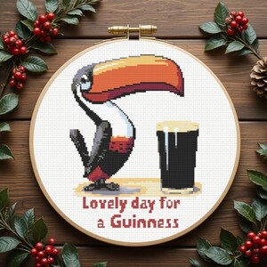 May include: A cross-stitch hoop art piece featuring a toucan and a pint of Guinness. The toucan has a large orange and black beak, with the text "Lovely day for a Guinness" below. The piece is set against a wooden background with festive greenery.