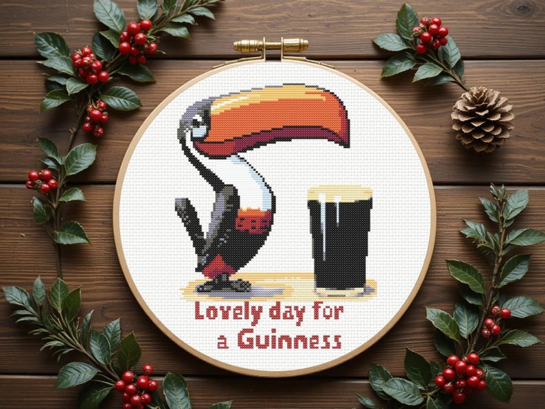 May include: A cross-stitch hoop art piece featuring a toucan and a pint of Guinness. The toucan has a large orange and black beak, with the text "Lovely day for a Guinness" below. The piece is set against a wooden background with festive greenery.