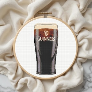 Pu&ograve; includere: Ricamo a punto croce di una pinta di Guinness stout in un bicchiere, con il logo Guinness. Il bicchiere &egrave; riempito di birra scura e una schiuma cremosa. Il ricamo &egrave; posto in un telaio di legno su sfondo bianco.