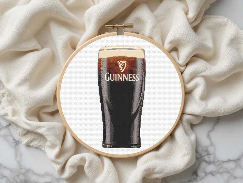Pu&ograve; includere: Ricamo a punto croce di una pinta di Guinness stout in un bicchiere, con il logo Guinness. Il bicchiere &egrave; riempito di birra scura e una schiuma cremosa. Il ricamo &egrave; posto in un telaio di legno su sfondo bianco.