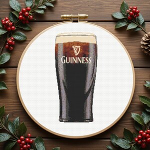 Pu&ograve; includere: Un disegno a punto croce con una pinta di Guinness. Il bicchiere &egrave; pieno di birra scura e una schiuma cremosa, con il logo Guinness visibile. Il disegno &egrave; ambientato in un telaio da ricamo in legno, circondato da verde e bacche rosse.