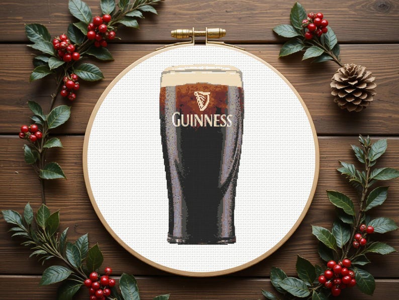 Pu&ograve; includere: Un disegno a punto croce con una pinta di Guinness. Il bicchiere &egrave; pieno di birra scura e una schiuma cremosa, con il logo Guinness visibile. Il disegno &egrave; ambientato in un telaio da ricamo in legno, circondato da verde e bacche rosse.