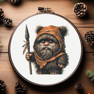 Könnte beinhalten: Kreuzstich eines Ewok-Charakters mit einer orangefarbenen Kapuze und einem Speer. Der Kreuzstich befindet sich in einem runden Holzreifen, umgeben von Tannenzapfen und Grün auf einer Holzoberfläche. Der Ewok hat ein detailliertes, pelziges Aussehen.