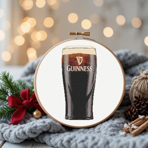 Pu&ograve; includere: Un disegno a punto croce di una pinta di Guinness in un telaio di legno. La birra scura ha una schiuma cremosa, con il logo Guinness. Lo sfondo include una coperta grigia, aghi di pino e decorazioni festive. Il telaio misura circa 20 cm di diametro.