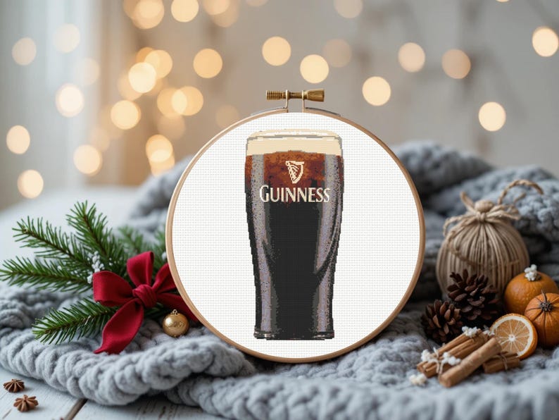 Pu&ograve; includere: Un disegno a punto croce di una pinta di Guinness in un telaio di legno. La birra scura ha una schiuma cremosa, con il logo Guinness. Lo sfondo include una coperta grigia, aghi di pino e decorazioni festive. Il telaio misura circa 20 cm di diametro.