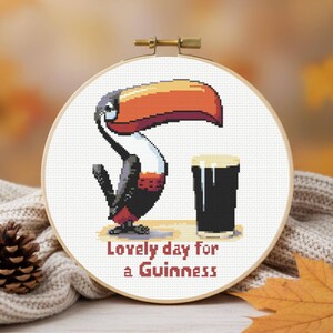 May include: Cross-stitch embroidery in a wooden hoop features a toucan with a large orange and red beak, a pint of stout, and the text "Lovely day for a Guinness". The artwork is set against a blurred background.