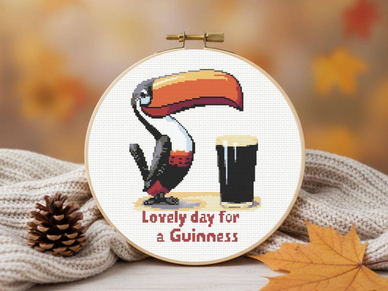 May include: Cross-stitch embroidery in a wooden hoop features a toucan with a large orange and red beak, a pint of stout, and the text "Lovely day for a Guinness". The artwork is set against a blurred background.