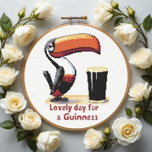 May include: Cross-stitch embroidery in a wooden hoop featuring a pixelated toucan with a large orange beak and a pint of Guinness. The text "Lovely day for a Guinness" is stitched below. White roses surround the hoop.