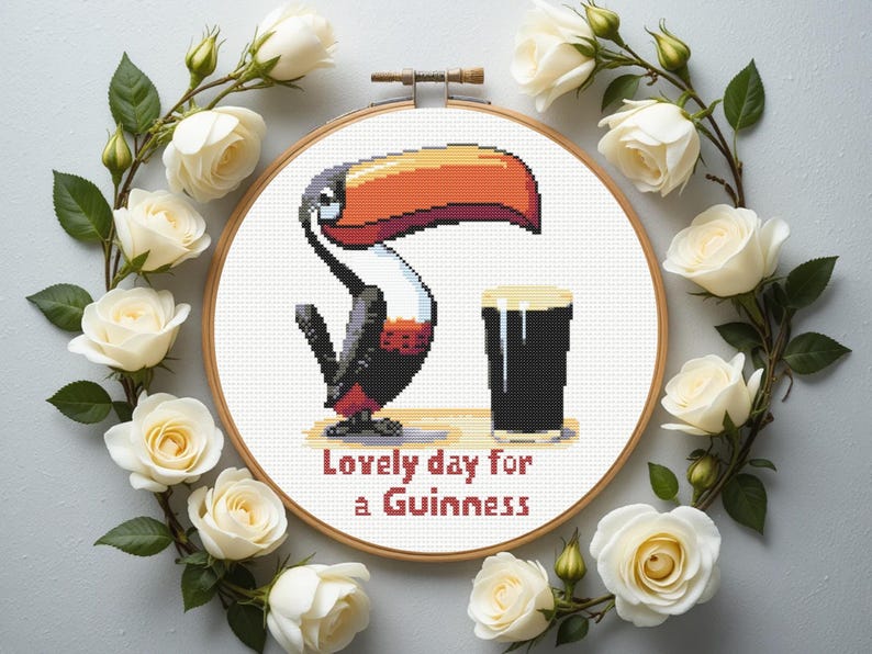 May include: Cross-stitch embroidery in a wooden hoop featuring a pixelated toucan with a large orange beak and a pint of Guinness. The text "Lovely day for a Guinness" is stitched below. White roses surround the hoop.