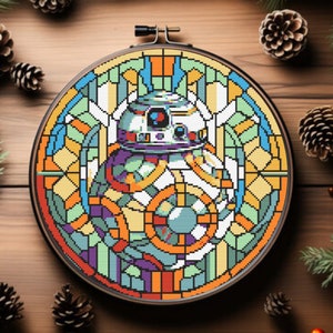 May include: A colorful cross-stitch embroidery of a BB-8 droid in a stained-glass style, set in a round wooden hoop. The design features geometric shapes in shades of orange, blue, green, and purple. The background includes pinecones and greenery.