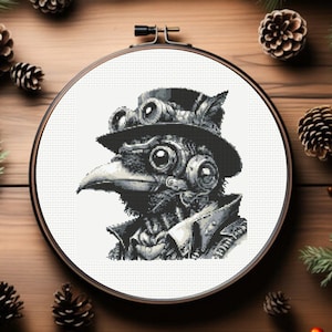 Puede incluir: Un diseño de punto de cruz en blanco y negro de un cuervo steampunk con sombrero y gafas, en un bastidor de bordado. El diseño está rodeado de agujas de pino, piñas y pequeñas bayas naranjas sobre una superficie de madera.