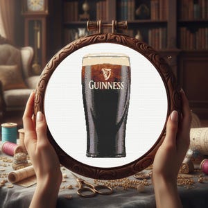 Pu&ograve; includere: Ricamo a punto croce di una pinta di Guinness in un telaio di legno. La birra scura ha una schiuma cremosa e il logo Guinness &egrave; visibile. Il telaio &egrave; tenuto davanti a un tavolo con materiale da cucito.