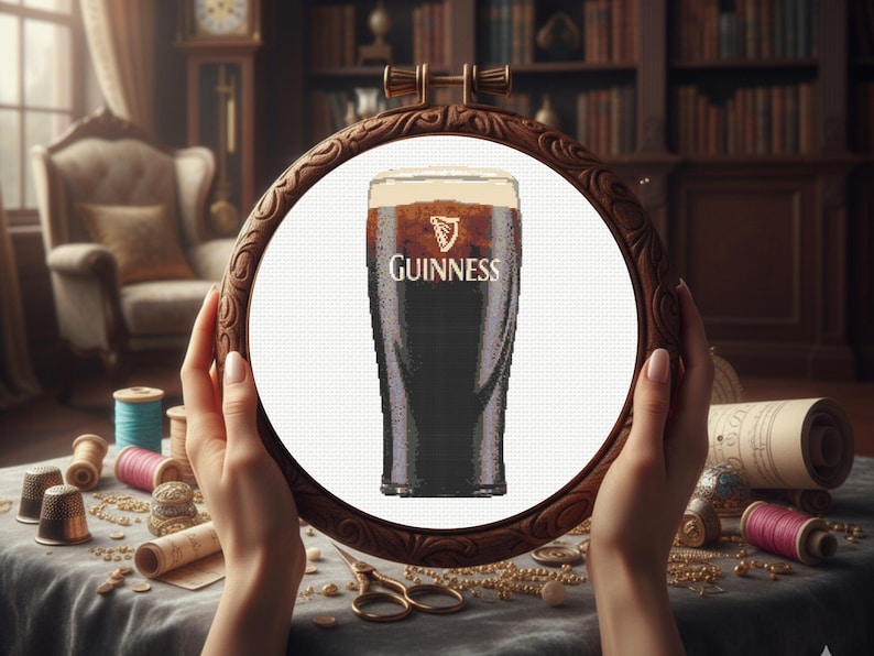 Pu&ograve; includere: Ricamo a punto croce di una pinta di Guinness in un telaio di legno. La birra scura ha una schiuma cremosa e il logo Guinness &egrave; visibile. Il telaio &egrave; tenuto davanti a un tavolo con materiale da cucito.