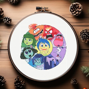 Può includere: Un telaio da ricamo a punto croce con i personaggi del film d'animazione "Inside Out". Il disegno include Gioia, Tristezza, Rabbia, Paura, Disgusto e altri personaggi. Il telaio è ambientato su uno sfondo di legno con pigne e verde.