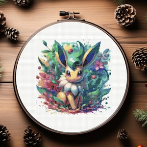 May include: A cross-stitch embroidery hoop featuring a colourful image of an Eevee Pokemon surrounded by flowers and leaves. The background is a watercolour-style wash of green, blue, and purple.