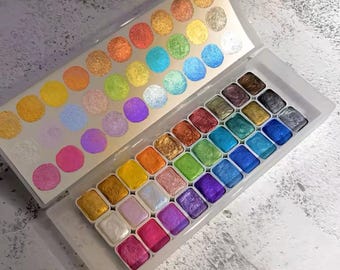 Puede incluir: Un juego de 36 acuarelas en una caja de plástico blanca. Las pinturas están dispuestas en filas y columnas, y cada pintura tiene un color diferente. Los colores son principalmente metálicos e iridiscentes, con algunos colores sólidos. La caja tiene una tapa transparente que muestra los colores de las pinturas.