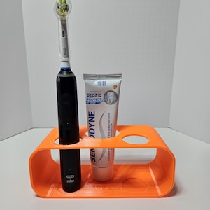 May include: An orange toothbrush holder with a black electric toothbrush and a tube of Sensodyne toothpaste. The toothpaste tube is white with blue and orange text. The toothbrush has a green and yellow brush head.