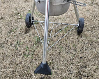 May include: A silver and brown charcoal grill with three legs and black wheels. The grill is on a patch of dry grass.