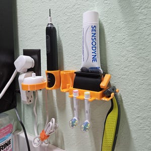 May include: A 3D printed orange and black bathroom organizer with a toothbrush holder, a razor holder, and a toothpaste dispenser. The organizer is mounted on a white tiled wall. The toothpaste tube is labeled "Sensodyne".
