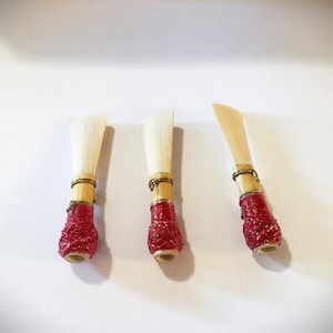 May include: Three wooden oboe reeds with red and gold accents. The reeds are tied together with a thin gold wire.