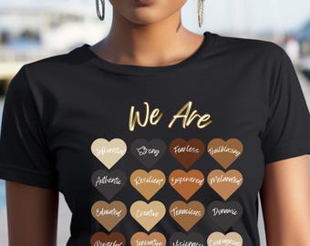 Pode incluir: Camiseta preta com o texto "We Are" em letras douradas. A camiseta apresenta uma grade de 12 corações, cada um de um tom de marrom diferente, com uma palavra descrevendo uma qualidade positiva escrita em branco dentro de cada coração. As palavras são: Influential, Strong, Fearless, Trailblazing, Authentic, Resilient, Empowered, Melanin, Educated, Creative, Tenacious, Dynamic, Powerful, Innovative, Visionary, Courageous.