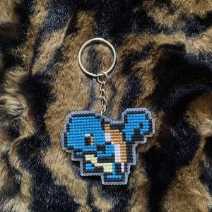 Puede incluir: Un llavero pixelado azul y blanco con un personaje de dibujos animados. El llavero está unido a un anillo y una cadena de color dorado. El fondo es un estampado de animales marrón y negro.