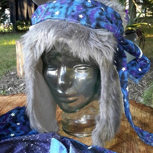 May include: A gray faux fur hat with blue and purple galaxy print fabric and white stars. The hat has long ear flaps and a matching scarf.
