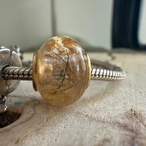 May include: A clear, round bead with a golden hue, containing delicate black inclusions. The bead is set with gold-colored metal accents and is strung on a silver-toned bracelet. The bead is approximately 0.75 inches in diameter.