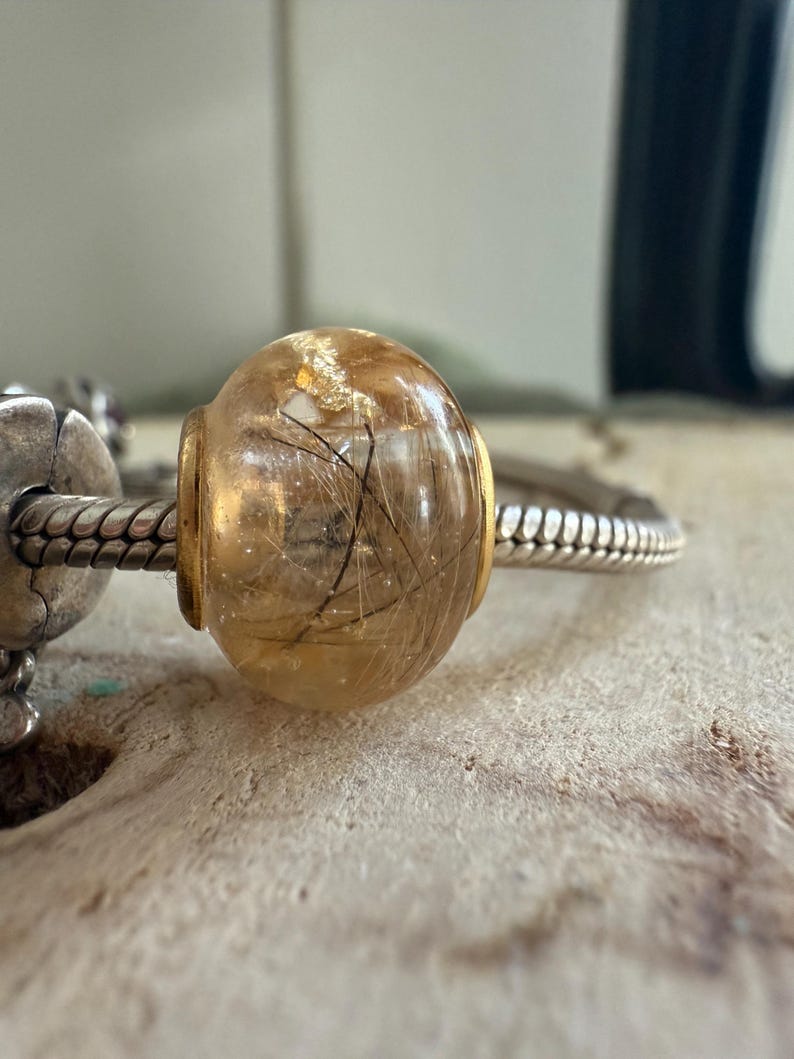 May include: A clear, round bead with a golden hue, containing delicate black inclusions. The bead is set with gold-colored metal accents and is strung on a silver-toned bracelet. The bead is approximately 0.75 inches in diameter.