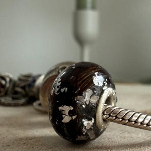 May include: Close-up of a dark brown bead with silver foil accents, strung on a silver chain. The bead has a rounded shape and a metallic sheen. Other beads are visible in the background, suggesting a jewelry theme.