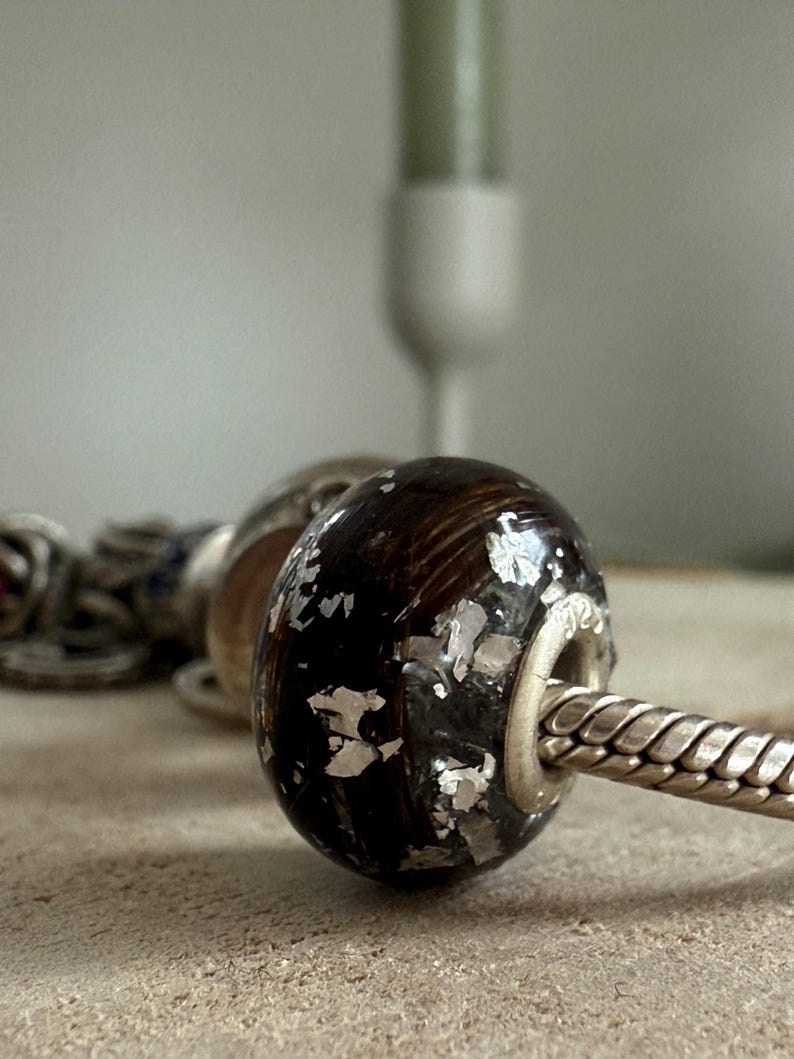 May include: Close-up of a dark brown bead with silver foil accents, strung on a silver chain. The bead has a rounded shape and a metallic sheen. Other beads are visible in the background, suggesting a jewelry theme.