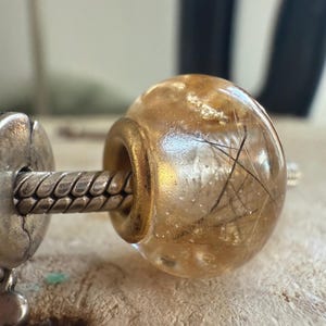 May include: A clear, round bead with a gold-colored metal ring. The bead contains delicate, dark-colored inclusions, and is attached to a silver-colored metal chain. The bead is a decorative element.