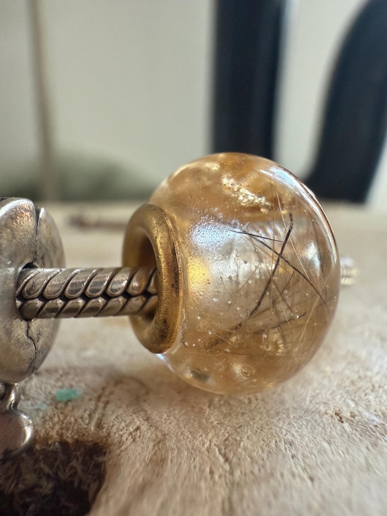 May include: A clear, round bead with a gold-colored metal ring. The bead contains delicate, dark-colored inclusions, and is attached to a silver-colored metal chain. The bead is a decorative element.