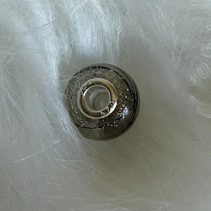 May include: A silver and gray bead with black lines and glitter accents. The bead has a silver ring attached. The bead is round and sits on a white, fluffy surface.