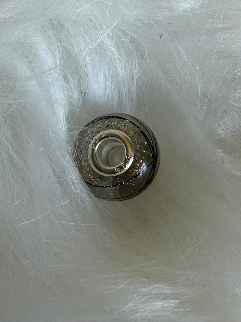 May include: A silver and gray bead with black lines and glitter accents. The bead has a silver ring attached. The bead is round and sits on a white, fluffy surface.
