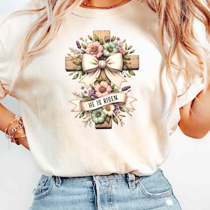 May include: A white t-shirt with a floral design featuring a wooden cross with a white bow and the text "He is Risen".