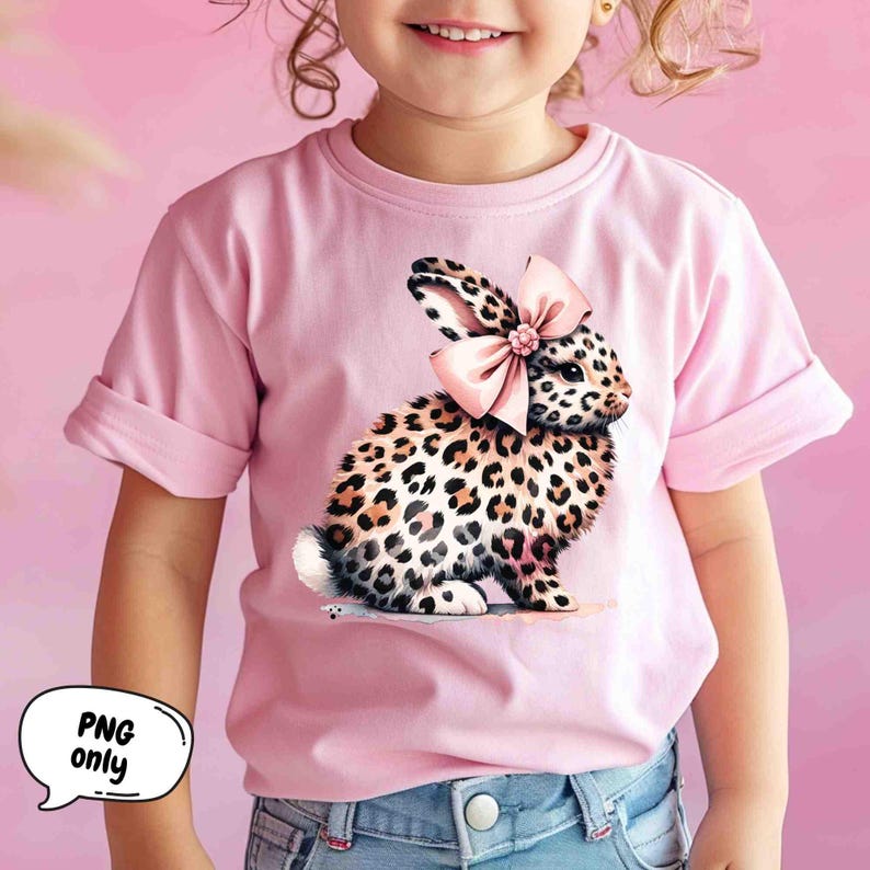 May include: A pink t-shirt with a leopard print bunny wearing a pink bow. The bunny is sitting on the shirt.