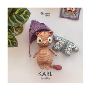 Könnte beinhalten: Eine gehäkelte Katzendoll namens Karl, mit rotem Schwanz und lila Hut. Die Puppe hat große weiße Augen und einen traurigen Ausdruck. Der Text "KARL the red Cat" steht darunter. Ein gehäkelter Pullover und ein Quadrat mit einem gehäkelten Katzengesicht sind ebenfalls zu sehen.