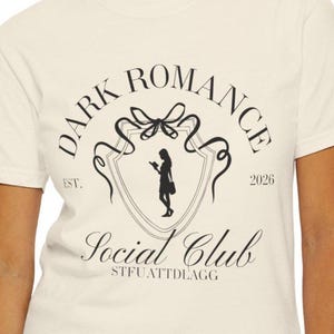 以下が含まれることがあります： クリーム色のTシャツに黒いグラフィックデザイン。デザインには「DARK ROMANCE SOCIAL CLUB」と年号「2026」の文字が含まれています。携帯電話を持っている人のシルエットがデザインの中央にあります。