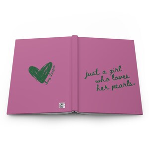 May include: Open journal with a magenta cover. The left side features a green heart and the words "Ivy League." The right side has the phrase "just a girl who loves her pearls" in green script. A small barcode is visible.
