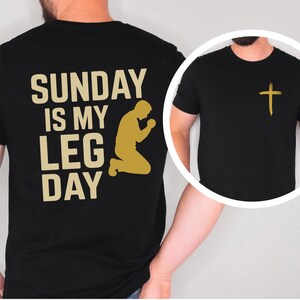 May include: Black t-shirt with a gold cross on the front and the text "SUNDAY IS MY LEG DAY" with a praying figure on the back. The text and figure are in a gold color.