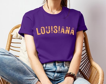 Puede incluir: Una camiseta morada con la palabra "LOUISIANA" impresa en naranja en el pecho. La camiseta tiene mangas cortas y cuello redondo. La modelo lleva vaqueros azules y está sentada en una silla de mimbre.