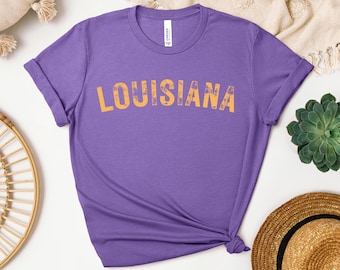 Puede incluir: Una camiseta morada con la palabra "LOUISIANA" impresa en naranja en el pecho. La camiseta tiene mangas cortas y cuello redondo. El texto tiene un aspecto desgastado y vintage.