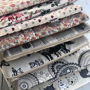 May include: A stack of eight fabric zippered pouches with different patterns. The patterns include floral, hearts, newspaper print, horses, mandalas, and sheep. The pouches are all beige with black accents.