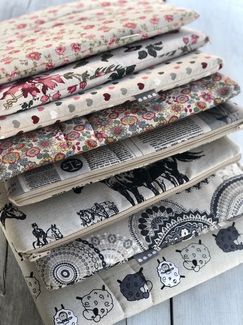 May include: A stack of eight fabric zippered pouches with different patterns. The patterns include floral, hearts, newspaper print, horses, mandalas, and sheep. The pouches are all beige with black accents.